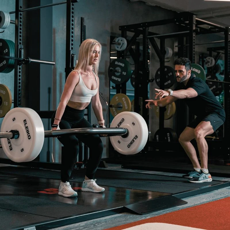Person lifting a barbell in a gym while a trainer provides guidance
